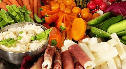 Colorful picture of cut peas, carrots, tomatoes, cucumbers, cheese, meat wrapped cheese and a dip.