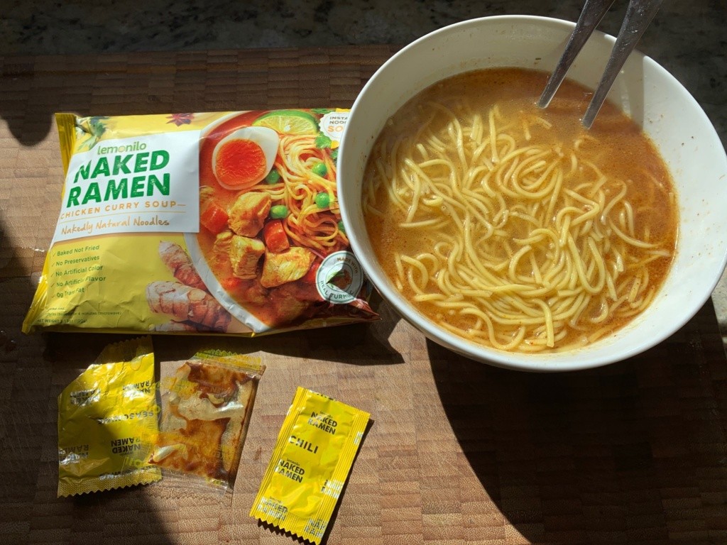 Photo of the noodle soup in a white bowl surrounded by the package and 4 seasoning packets.