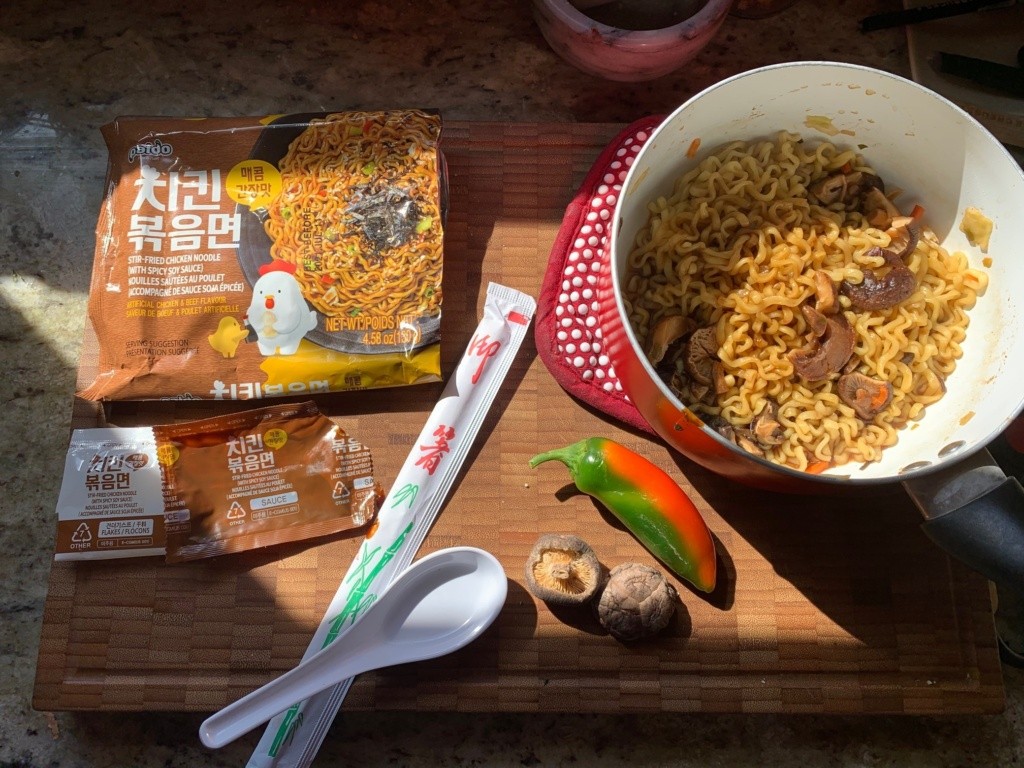 Photo of a pot of noodles with its packaging, chopsticks, spoon and dried mushrooms and a pepper.