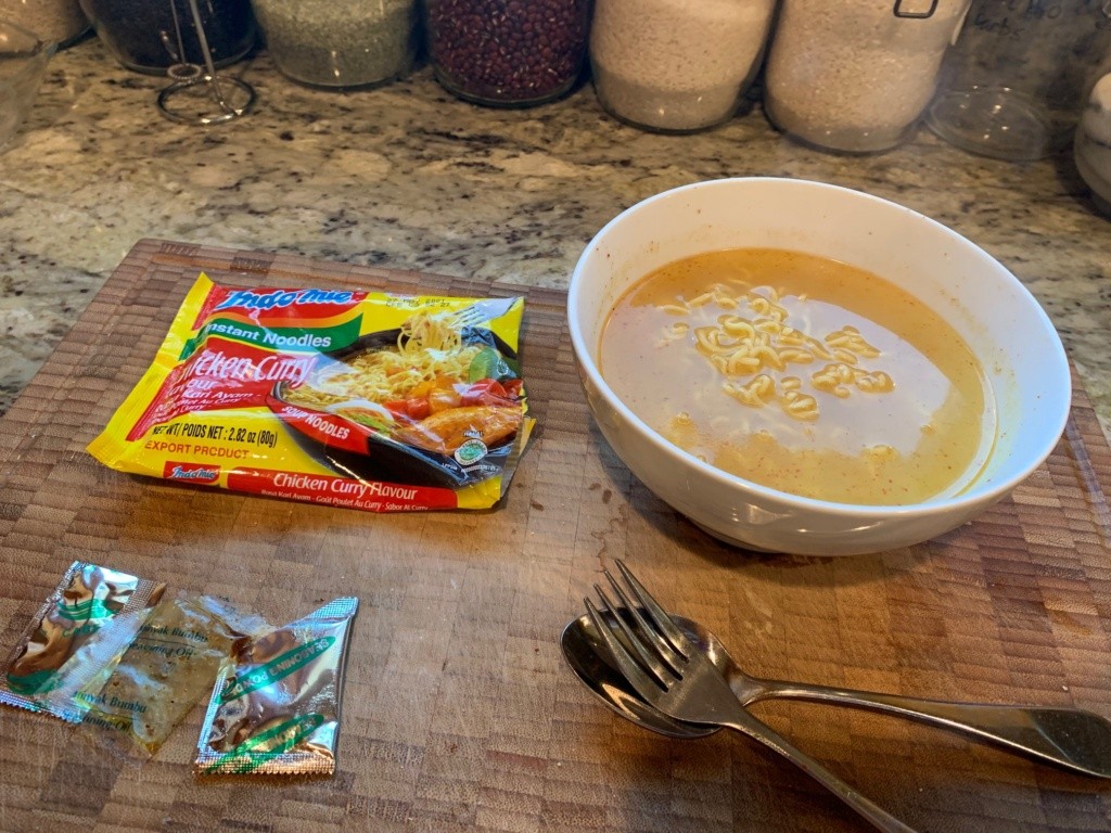 Photo of the package, a bowl of noodle soup and a fork and spoon. The broth color is yellow , indicative of chicken.