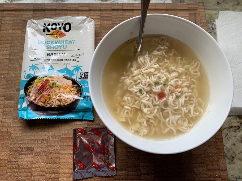 A picture of noodle soup and its packaging, with flecks of green, red, and orange veggies in the bowl.