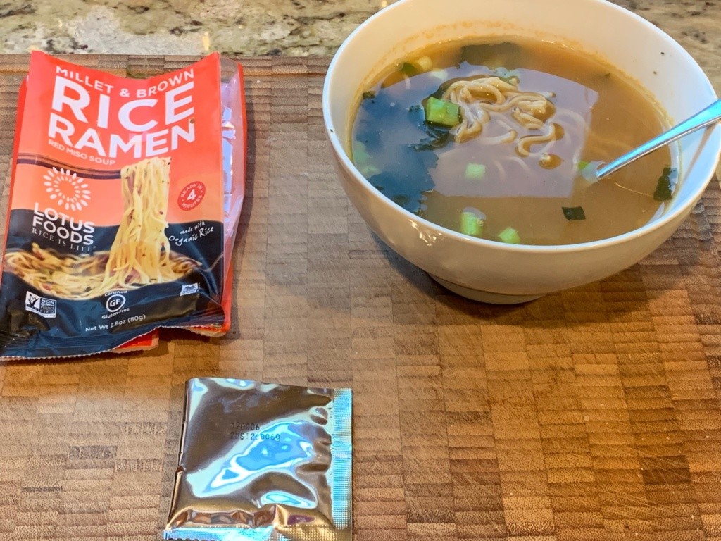 A brownish soup with seaweed and scallions in a white bowl with it packaging.