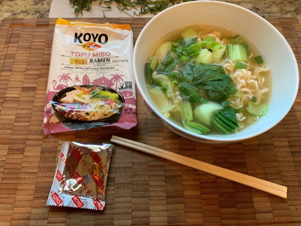 A picture of bowl of ramen soup and chopped bok choy, the packaging and chop sticks.