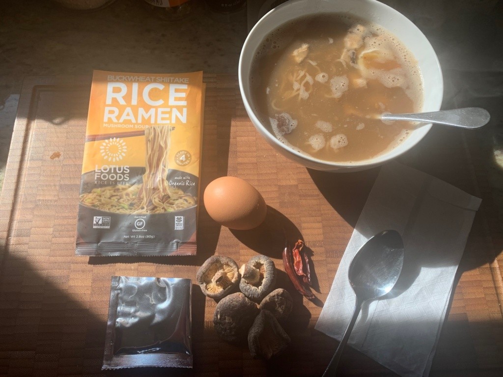 A muddy-brown mushroom broth soup in a white ceramic bowl and its packaging, and surrounded with an egg, mushrooms and chili peppers.