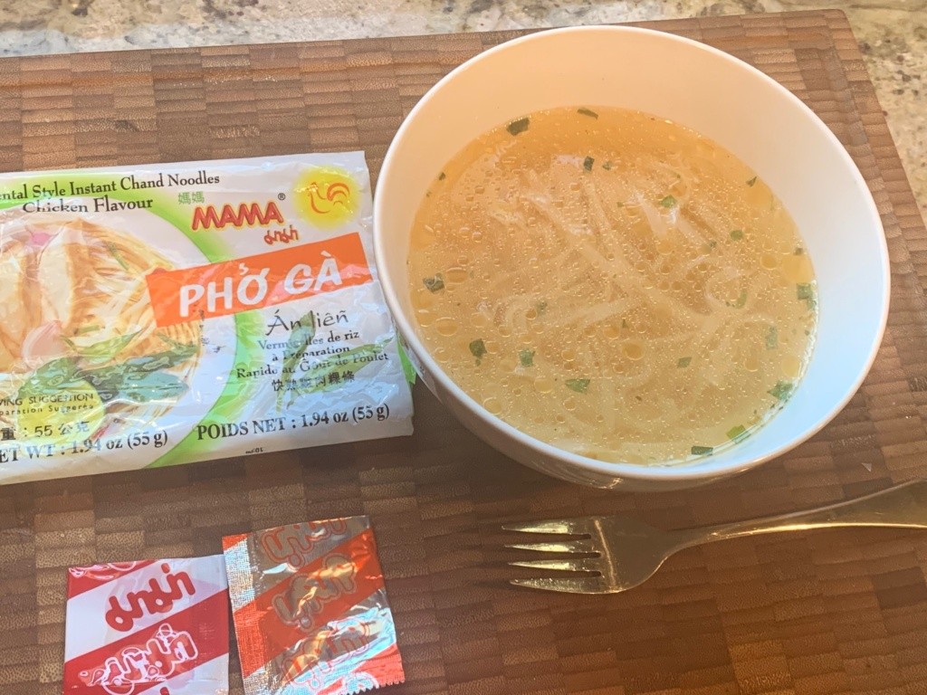 A bowl of Pho with packaging and for,. The soup is a light yellowish color with flecks of green.