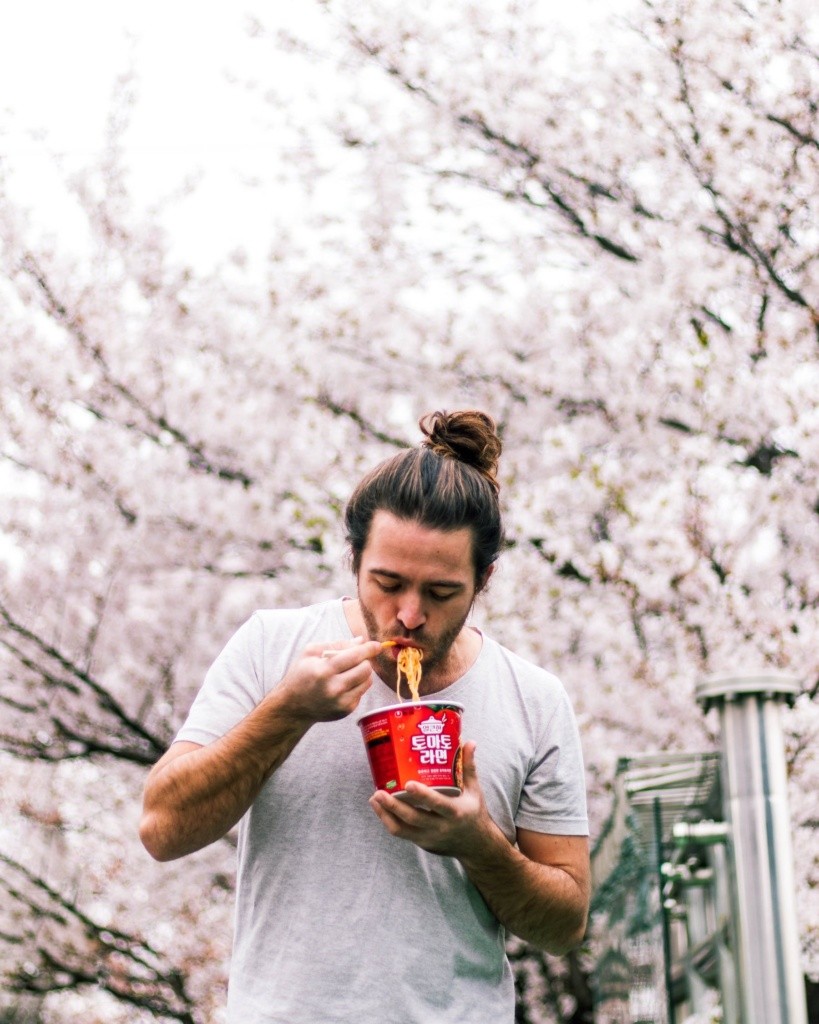 Photo of a man eating take out noodles and soup in from of a blooming cherry tree.